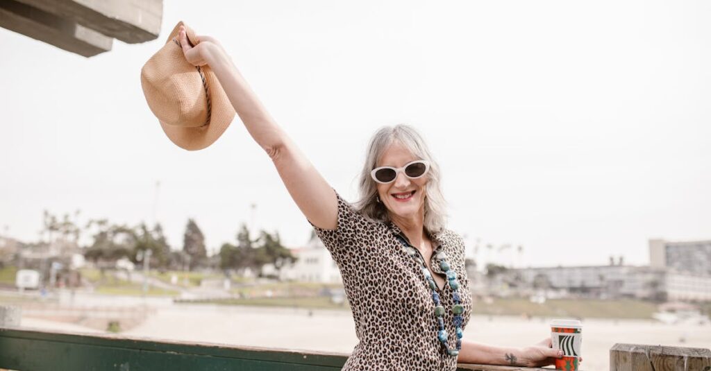Silver-haired woman in sunglasses smiling and raising a sun hat on a bright day at the beach.